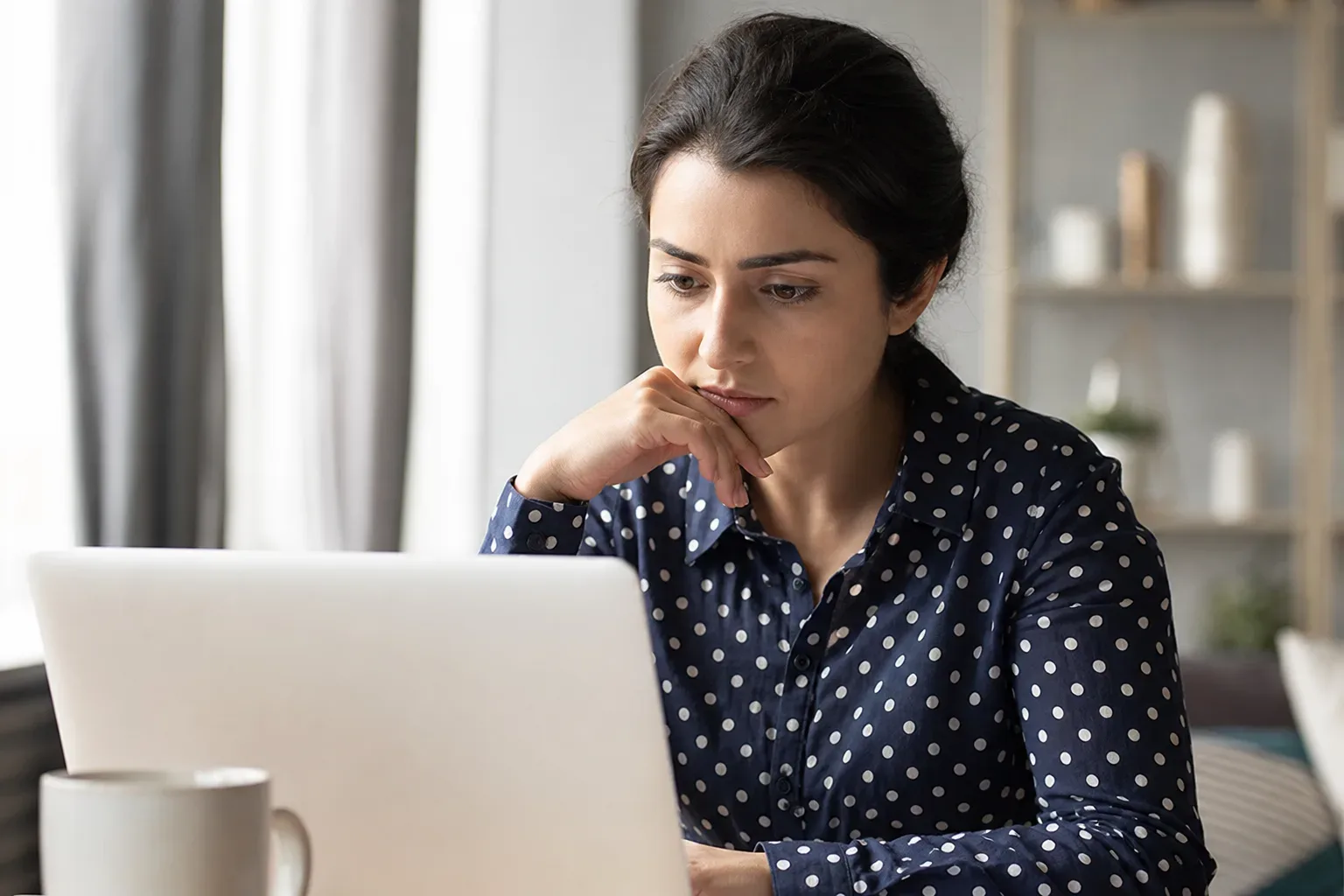 A person in a polka-dot shirt sits at a desk, resting their chin on their hand whilst focused on a laptop screen.