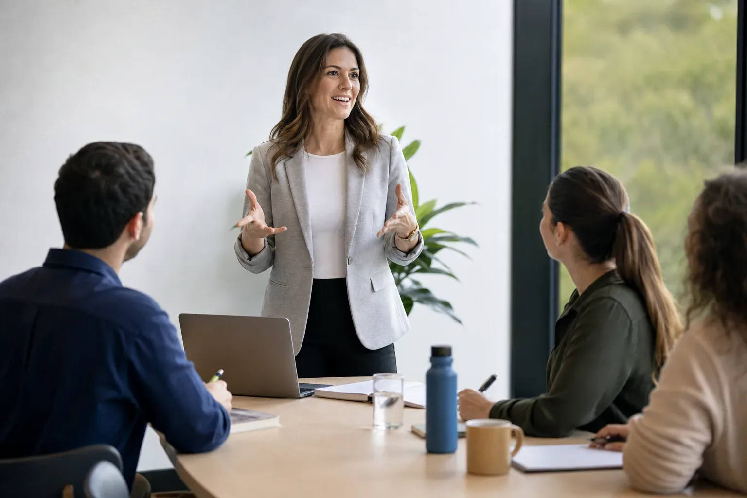A person in a grey blazer speaks animatedly to three colleagues seated around a table in a bright meeting room.