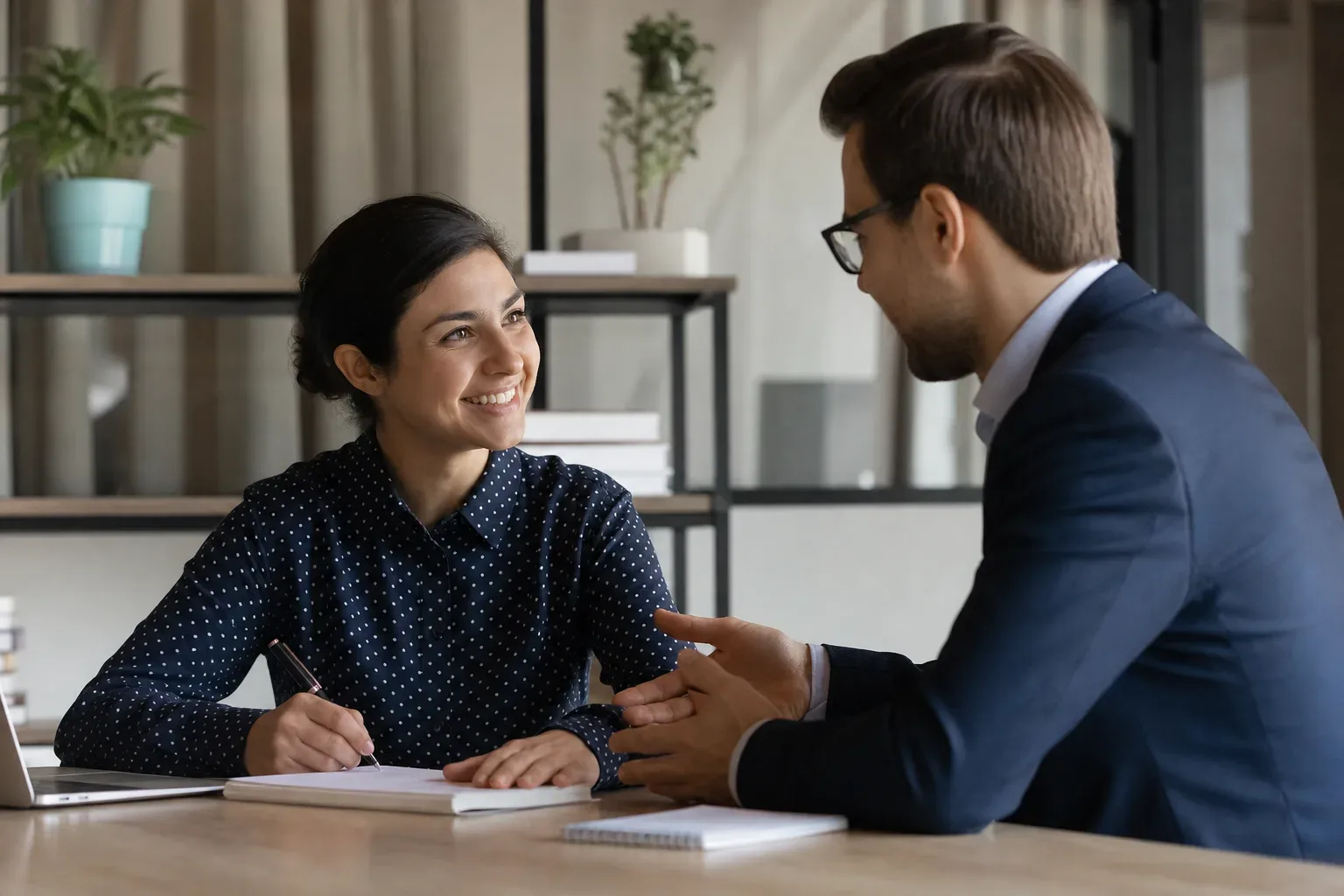 Two people sit at a desk in conversation, one taking notes and smiling, the other gesturing whilst speaking.