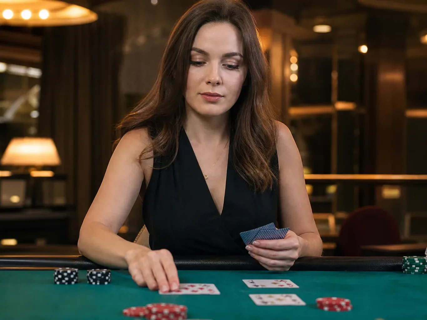 A person in a black dress deals cards at a green casino table, holding a hand of cards with poker chips nearby.