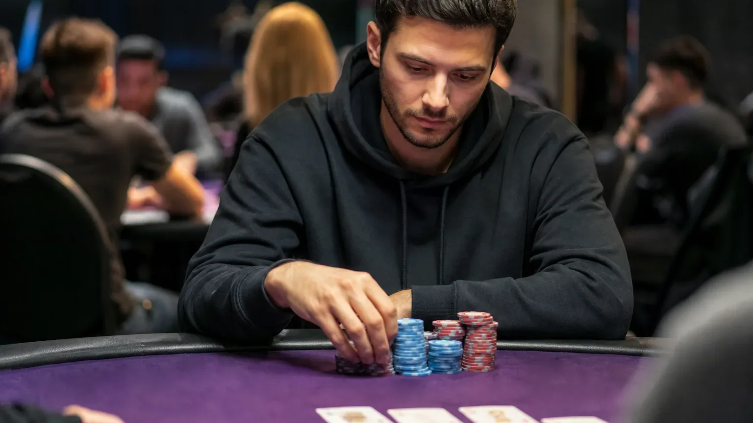 A person in a black hoodie stacks poker chips on a purple felt table during a casino card game.