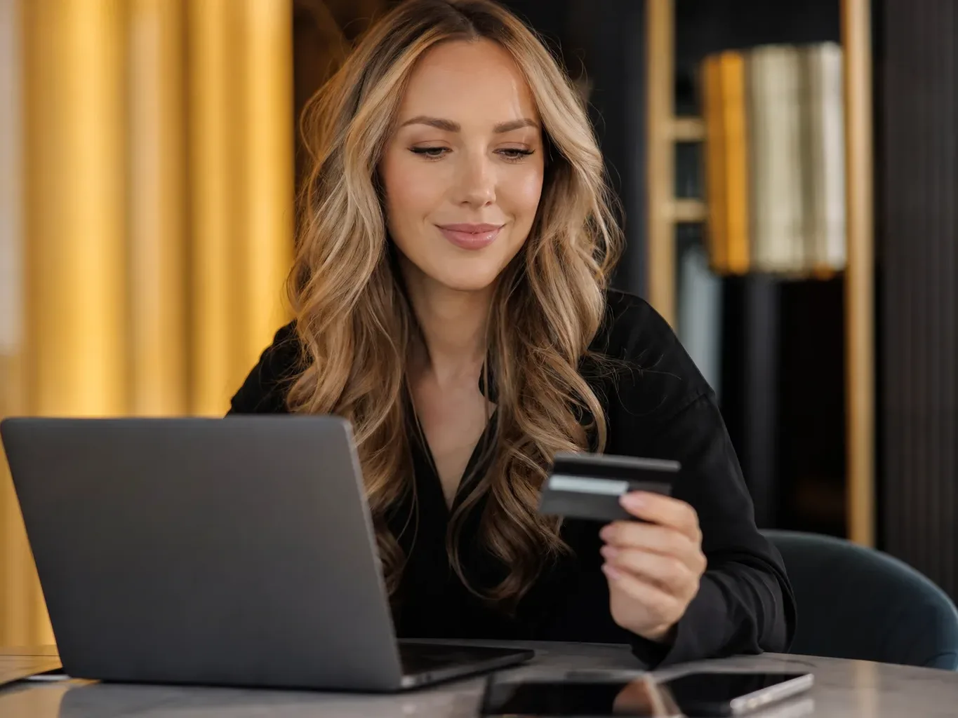 A person with long blonde hair holds a bank card whilst looking at an open laptop in a darkly furnished room.