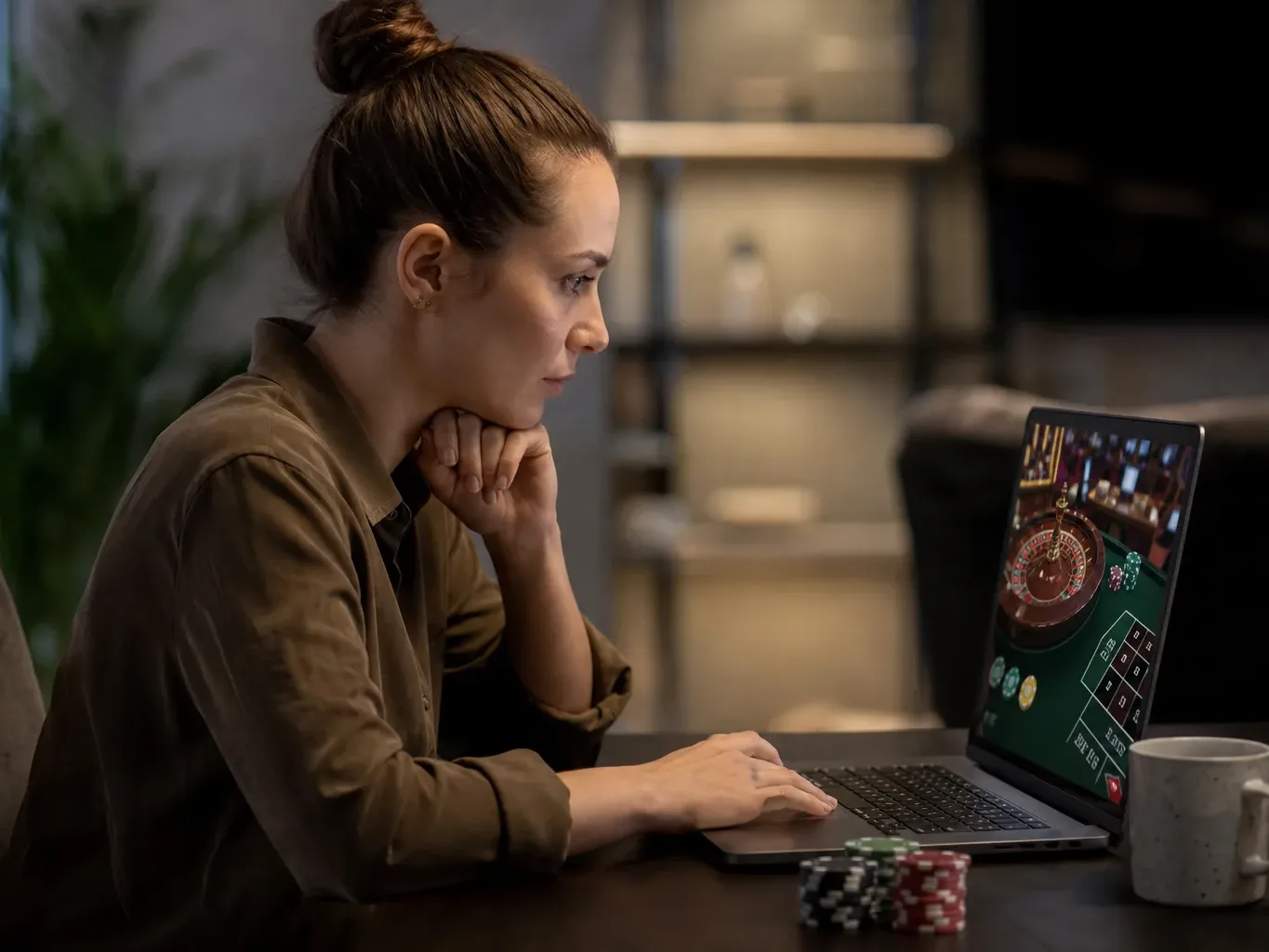 A person sits at a dark table at home, intently watching an online roulette game on a laptop, with poker chips nearby.