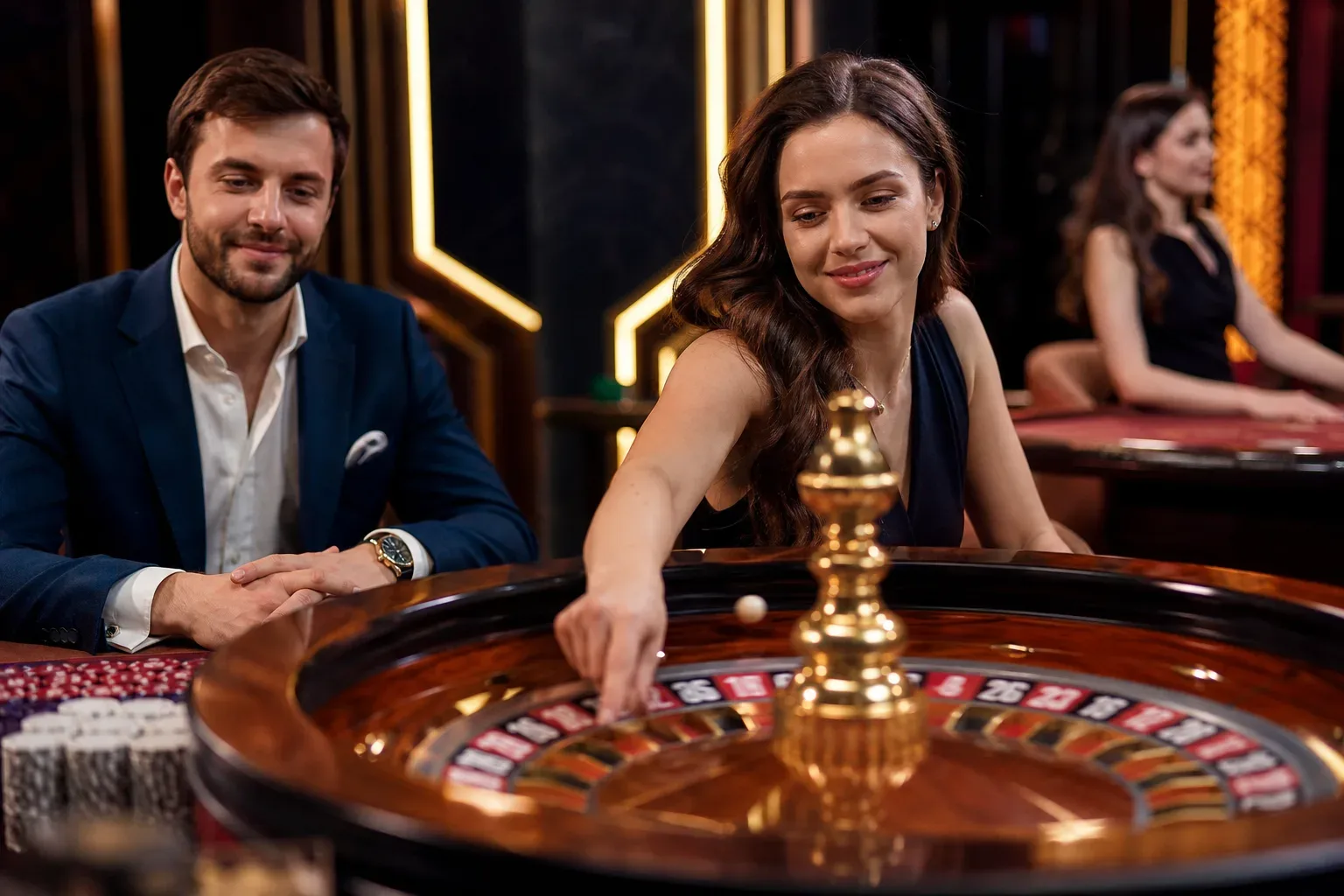 Two people lean over a roulette wheel at a casino table, smiling, with casino chips visible nearby.