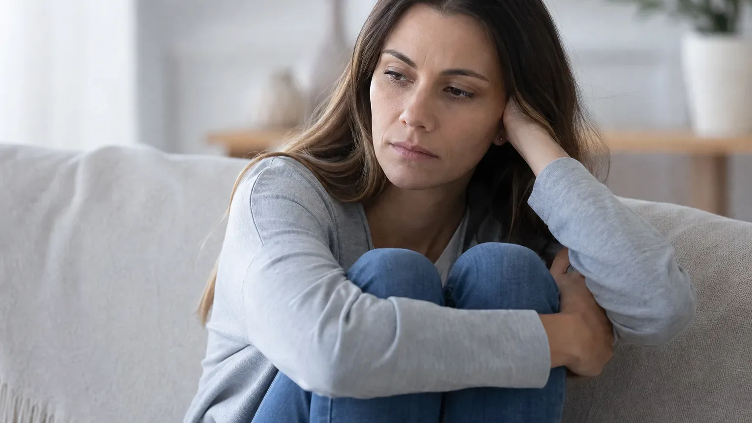 A person sits curled up on a sofa, hugging their knees and resting their head on one hand, looking downward with a troubled expression.